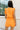 Back view of female model wearing the Raya Orange Denim Romper that has orange denim fabric, button up front, belt, and ruffle details.