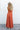 Full body back view of female model wearing the Jules Orange Printed Wide Leg Jumpsuit that has orange printed fabric, thin straps, and wide legs.