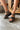 Frontal side view of female model wearing the Cienna Wedge Sandal in Black & Natural which features Black Faux Leather, Off White Stitch Detail, Criss-Cross Straps, Adjustable Back Strap, Lug Wedge Sole and Light Brown Cushion Details