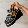 Side view of female model holding the Cienna Wedge Sandal in Black & Natural which features Black Faux Leather, Off White Stitch Detail,  Criss-Cross Straps, Adjustable Back Strap, Lug Wedge Sole and Light Brown Cushion Details
