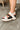 Side view of female model wearing the Joanie Slingback Wedge Sandal in Chalk & Black which features Ivory Suede and Black Leather Upper Fabric, Braid Details, Multi Straps, Adjustable Back Strap, Wedge Sole and 3 inch Heel Height