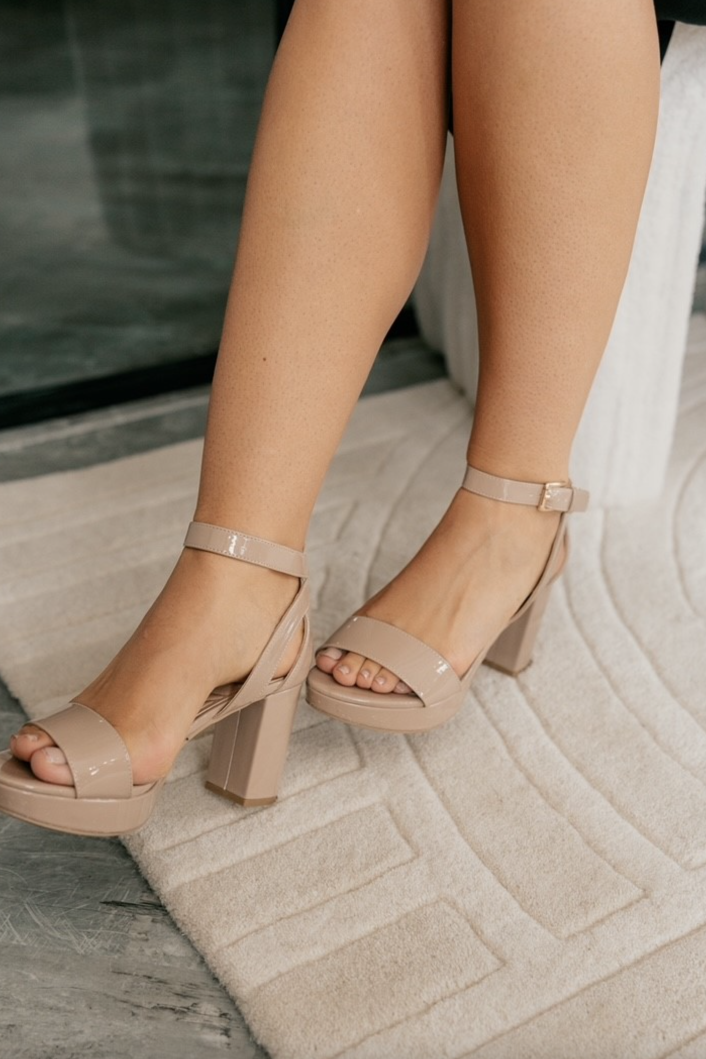 Right front angle view of female model wearing the Go On 2 Dress Sandal which features nude patent leather fabric, monochrome block heel, a delicate adjustable ankle strap, and a narrow vamp.