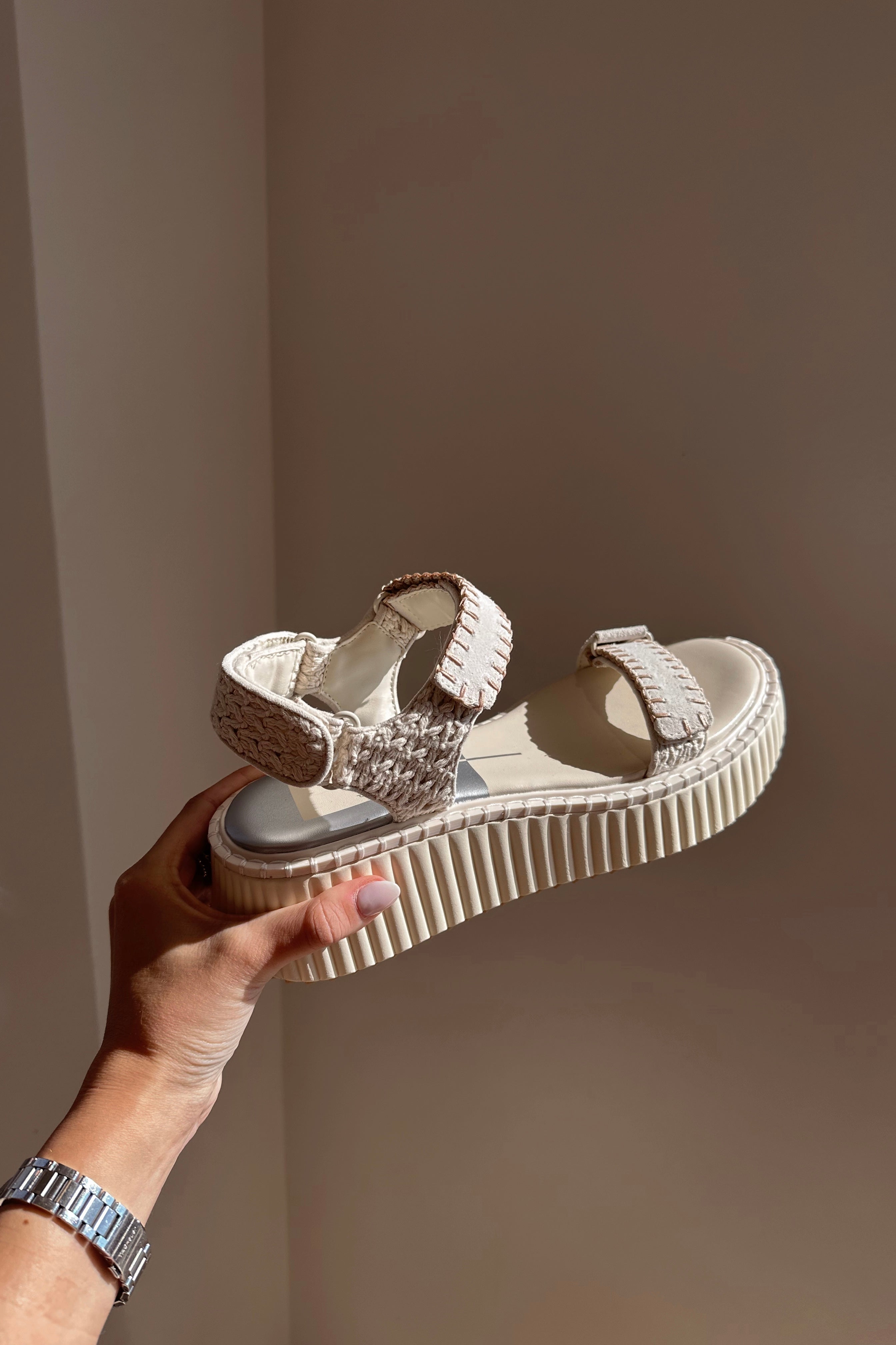 Side view of a hand holding the Debra Sandal in Ivory Suede which features ivory suede and white rattan fabric, white platform sole, rubber outsole and adjustable velcro straps