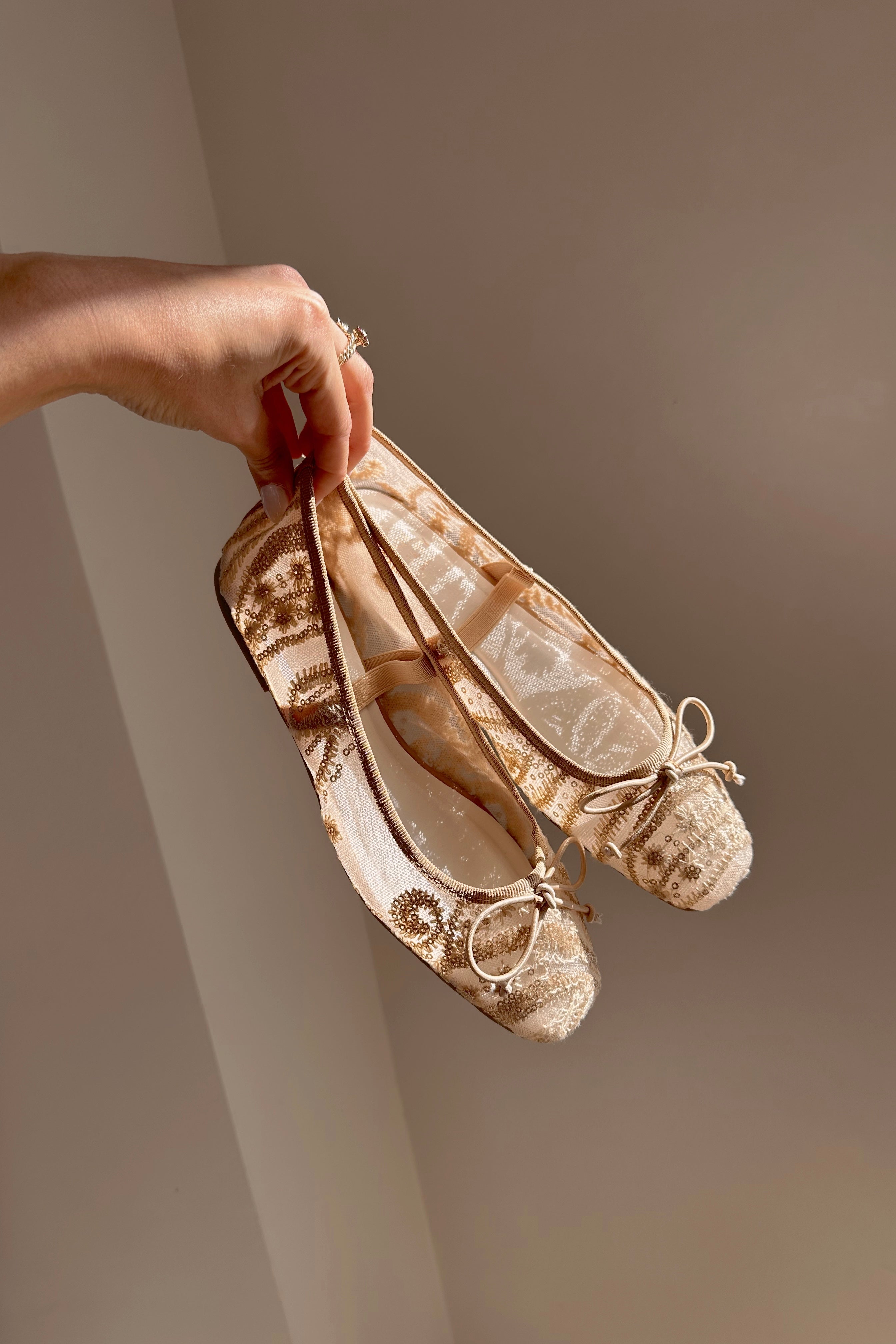 Side view of hand that is holding the Bisbee Ballet Flat in Gold that have beige mesh, ivory embroidery, gold sequins, a bow on front, and an elastic strap across the foot.
