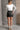 Full body back view of model wearing the Bexley Black Faux-Leather Mini Skirt that has black faux leather, white stitch details, a front zipper, two front pockets, and mini length.