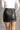Back view of model wearing the Bexley Black Faux-Leather Mini Skirt that has black faux leather, white stitch details, a front zipper, two front pockets, and mini length.