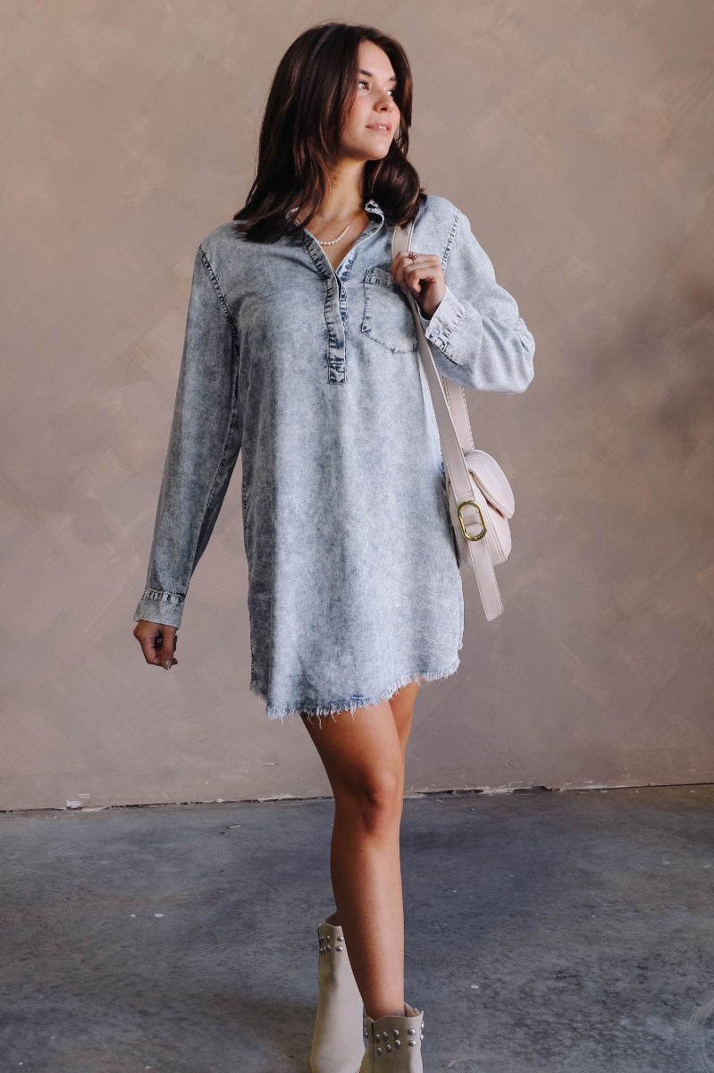 Full body front view of model wearing the Talk The Talk Dress In Blue that has washed blue denim fabric, mini length, a frayed hem, a hidden quarter button up, a collar, a chest pocket, and long sleeves.
