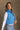 Side view of model wearing the Sutton Blue Cropped Puffer Vest that has blue puffer fabric, a  3 tiered design, medium blue lining, a cropped waist,pockets, a front zipper, a drawstring, a high neck and a sleeveless design.