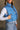 Side view of model wearing the Sutton Blue Cropped Puffer Vest that has blue puffer fabric, a  3 tiered design, medium blue lining, a cropped waist,pockets, a front zipper, a drawstring, a high neck and a sleeveless design.