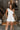 Front view of model wearing the All My Love Ruffle Romper which features white fabric, pockets on each side, a tie around the waist, one shoulder ruffle strap, and a monochromatic side zipper with a hook closure.