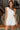 Front view of model wearing the All My Love Ruffle Romper which features white fabric, pockets on each side, a tie around the waist, one shoulder ruffle strap, and a monochromatic side zipper with a hook closure.