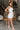 Full body side view of model wearing the All My Love Ruffle Romper which features white fabric, pockets on each side, a tie around the waist, one shoulder ruffle strap, and a monochromatic side zipper with a hook closure.