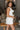 Side view of model wearing the All My Love Ruffle Romper which features white fabric, pockets on each side, a tie around the waist, one shoulder ruffle strap, and a monochromatic side zipper with a hook closure.