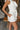 Close up side view of model wearing the All My Love Ruffle Romper which features white fabric, pockets on each side, a tie around the waist, one shoulder ruffle strap, and a monochromatic side zipper with a hook closure.