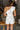 Back view of model wearing the All My Love Ruffle Romper which features white fabric, pockets on each side, a tie around the waist, one shoulder ruffle strap, and a monochromatic side zipper with a hook closure.