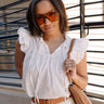 Front view of model wearing the Emory White Sleeveless Ruffle Top that features off white cotton fabric, a ruffled notched neckline with ties, and a sleeveless body with ruffle details. Shirt is styled tucked into pants with belt.