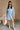 Full body view of model wearing the Melissa Light Denim Fray Mini Dress which features light denim tencel fabric, fray hem details, wooden button up front closure, two side slit pockets, mini length, v-neckline and long sleeves with buttoned cuffs.