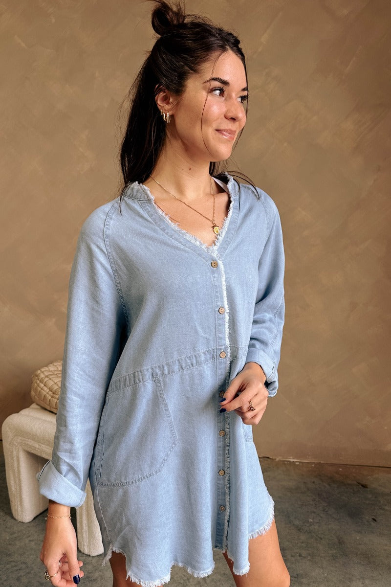 Front view of model wearing the Melissa Light Denim Fray Mini Dress which features light denim tencel fabric, fray hem details, wooden button up front closure, two side slit pockets, mini length, v-neckline and long sleeves with buttoned cuffs.