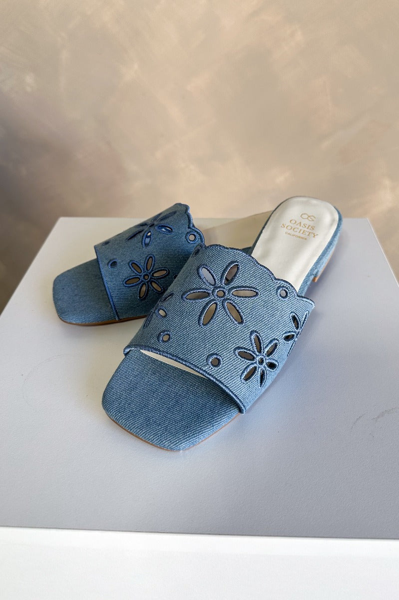 Front left angle view of the Manila Denim Eyelet Flower Heel Sandal which features blue denim fabric with eyelet flower details, square toes, backless slide entry, and 1" heels.