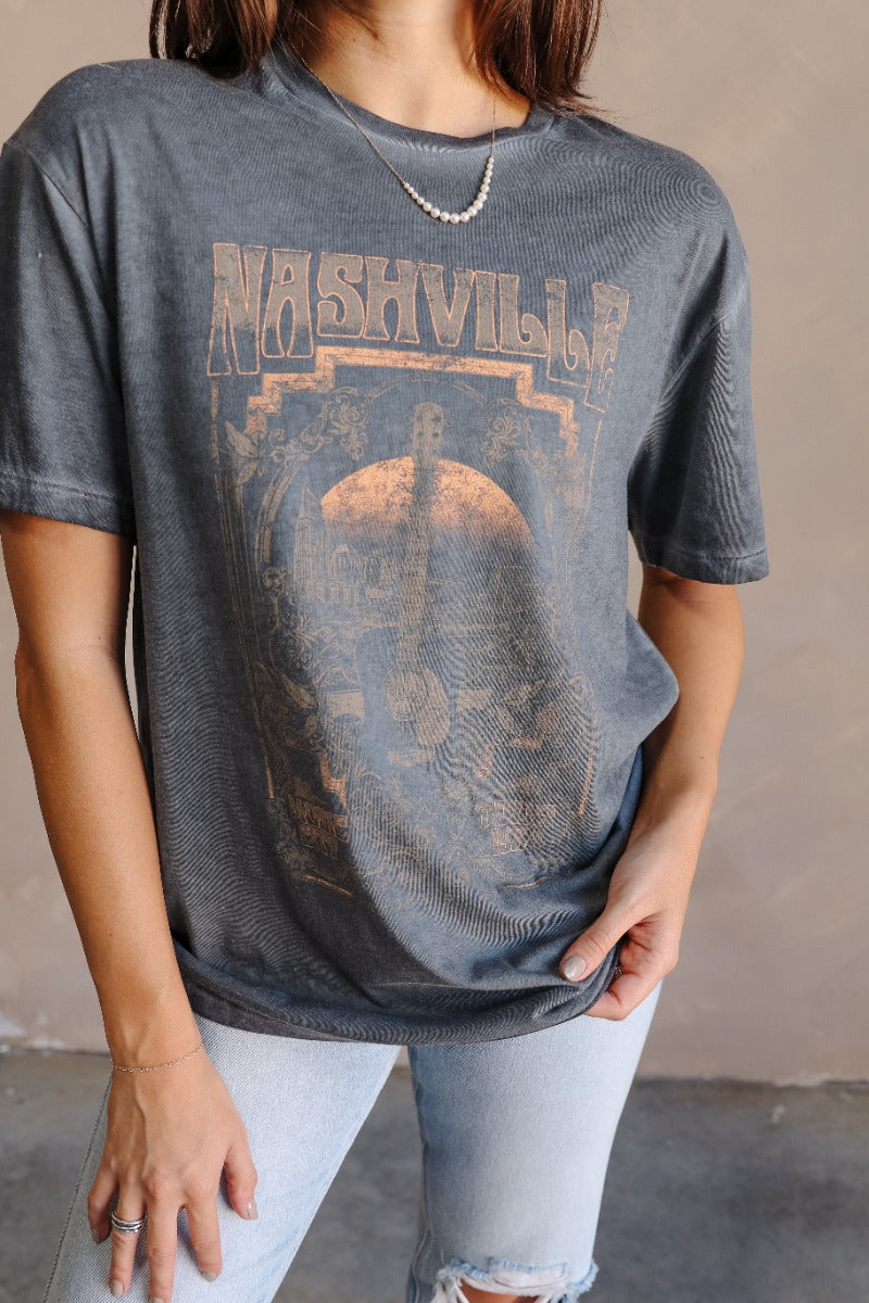 Close up view of model wearing the Nashville Charcoal Grey Short Sleeve Graphic Tee which features dark grey washed cotton fabric, round neckline and short sleeves. Graphic says "Nashville" with a guitar and music city scene graphic in light grey, rust, a