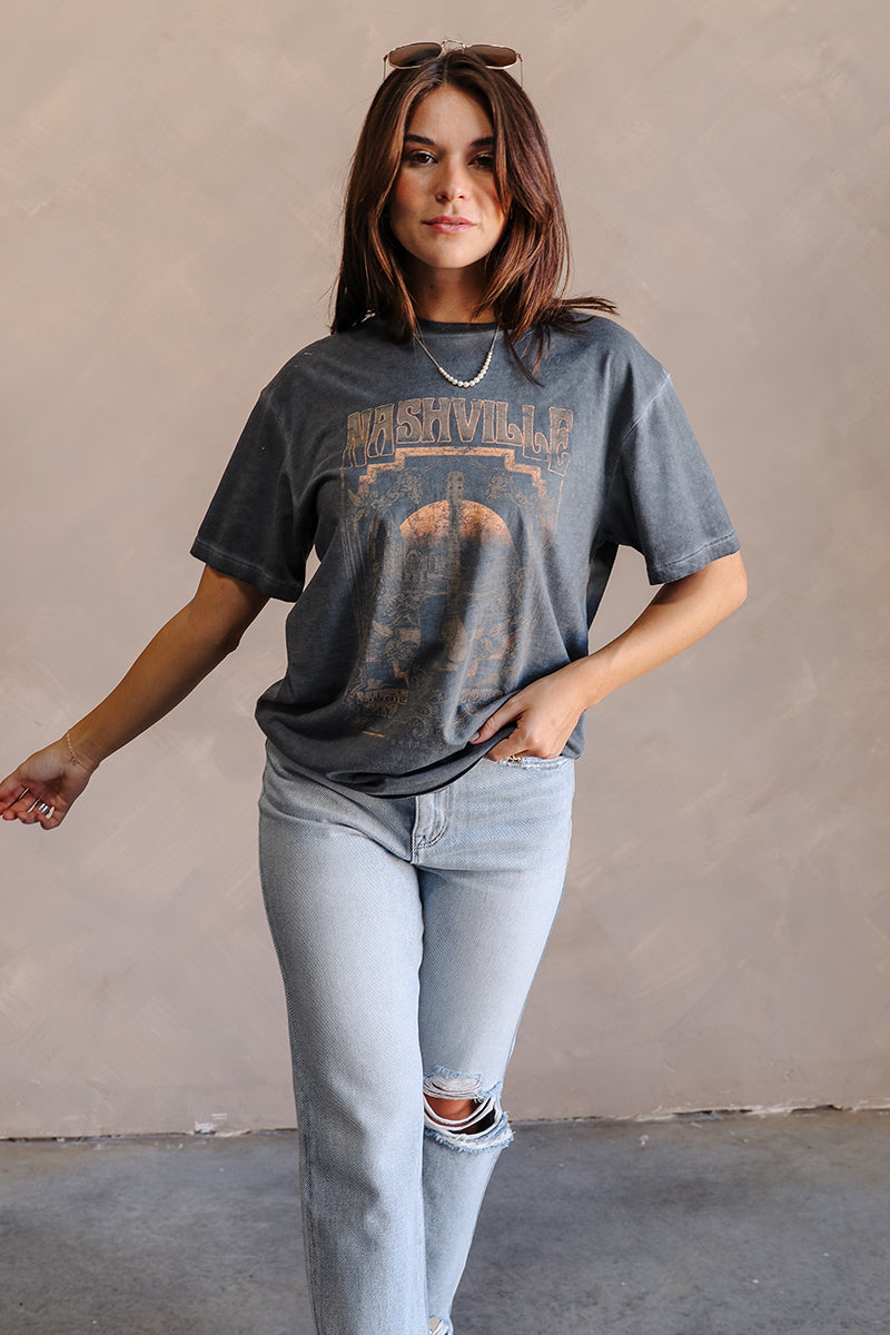 Full body view of model wearing the Nashville Charcoal Grey Short Sleeve Graphic Tee which features dark grey washed cotton fabric, round neckline and short sleeves. Graphic says "Nashville" with a guitar and music city scene graphic in light grey, rust, 