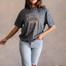 Full body view of model wearing the Nashville Charcoal Grey Short Sleeve Graphic Tee which features dark grey washed cotton fabric, round neckline and short sleeves. Graphic says "Nashville" with a guitar and music city scene graphic in light grey, rust, 