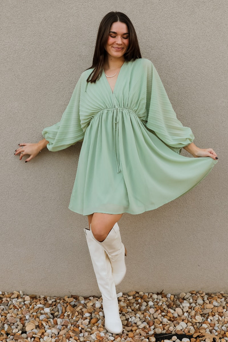Full body view of model wearing the Brooklyn Light Green Mini Dress which features light green fabric, mini length, light green lining, elastic waistband with drawstring ties, upper plisse fabric, a plunge neckline with a hook closure, 3/4 sleeves with el
