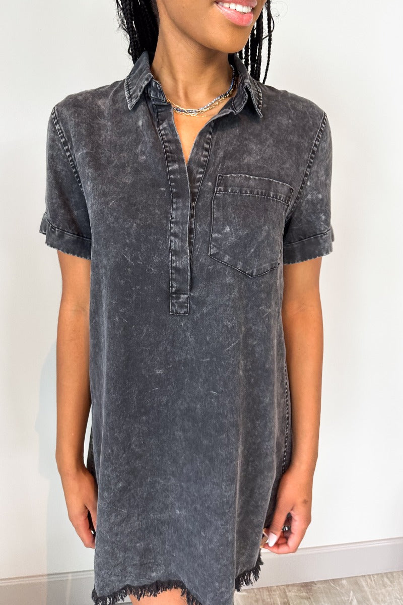 Close up view of model wearing the Play It Cool Dress in Washed Black which features washed light black fabric, one front chest pocket, a hidden quarter button-up, a v-neckline with a collar, a frayed mini-length hem, and cuffed short sleeves.