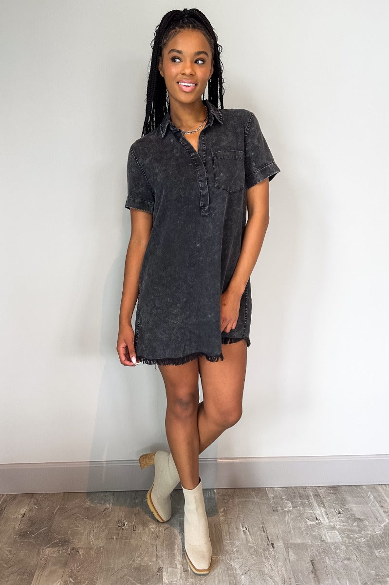 Full body view of model wearing the Play It Cool Dress in Washed Black which features washed light black fabric, one front chest pocket, a hidden quarter button-up, a v-neckline with a collar, a frayed mini-length hem, and cuffed short sleeves.