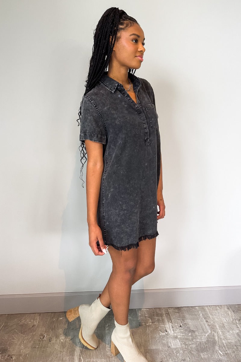 Full body side view of model wearing the Play It Cool Dress in Washed Black which features washed light black fabric, one front chest pocket, a hidden quarter button-up, a v-neckline with a collar, a frayed mini-length hem, and cuffed short sleeves.