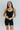 Front view of model wearing the Can You Keep Up Bodysuit that features black stretch fabric, a round neckline, adjustable thick straps, and fitted shorts.