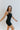 Side view of model wearing the Can You Keep Up Bodysuit that features black stretch fabric, a round neckline, adjustable thick straps, and fitted shorts.
