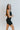 Side view of model wearing the Can You Keep Up Bodysuit that features black stretch fabric, a round neckline, adjustable thick straps, and fitted shorts.