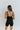 Back  view of model wearing the Can You Keep Up Bodysuit that features black stretch fabric, a round neckline, adjustable thick straps, and fitted shorts.