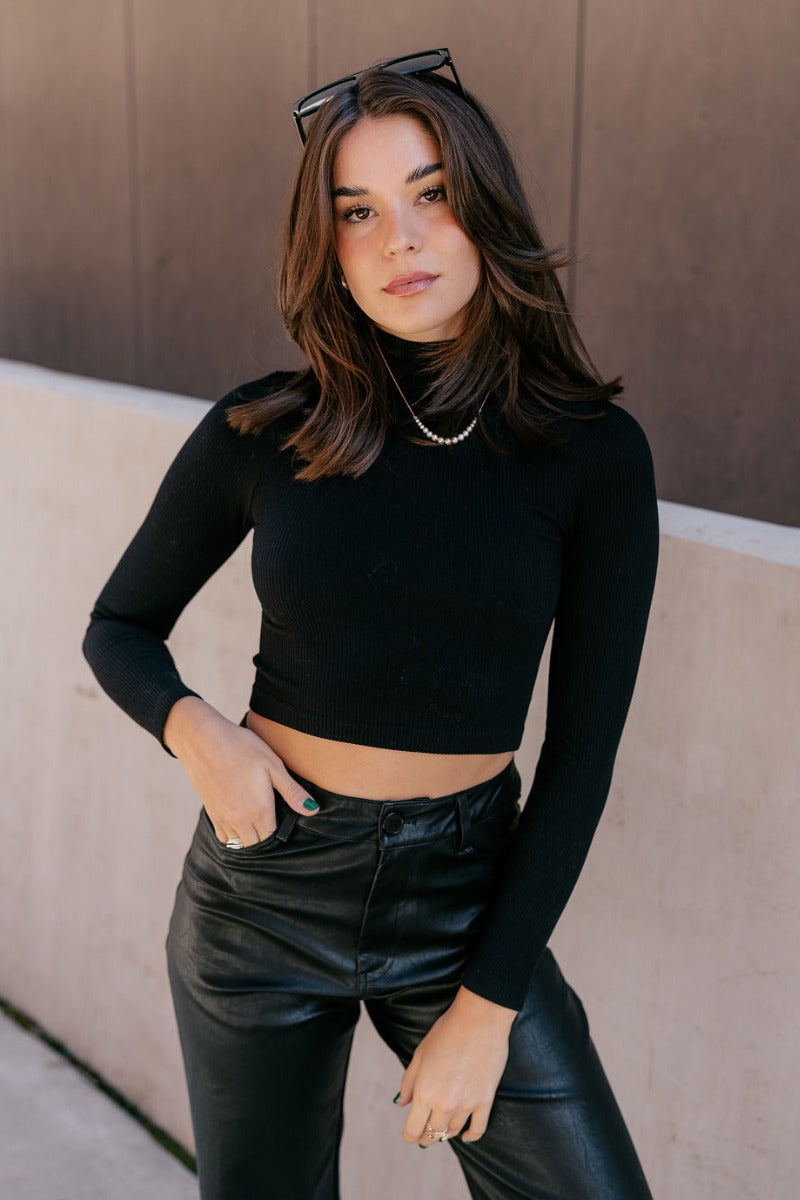 Front view of model wearing the Naomi Black High Neck Long Sleeve Top which features black ribbed fabric, a v-neckline, and long sleeves.