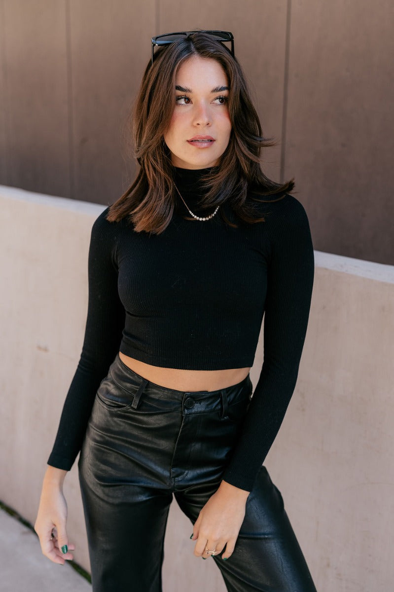 Front view of model wearing the Naomi Black High Neck Long Sleeve Top which features black ribbed fabric, a v-neckline, and long sleeves.