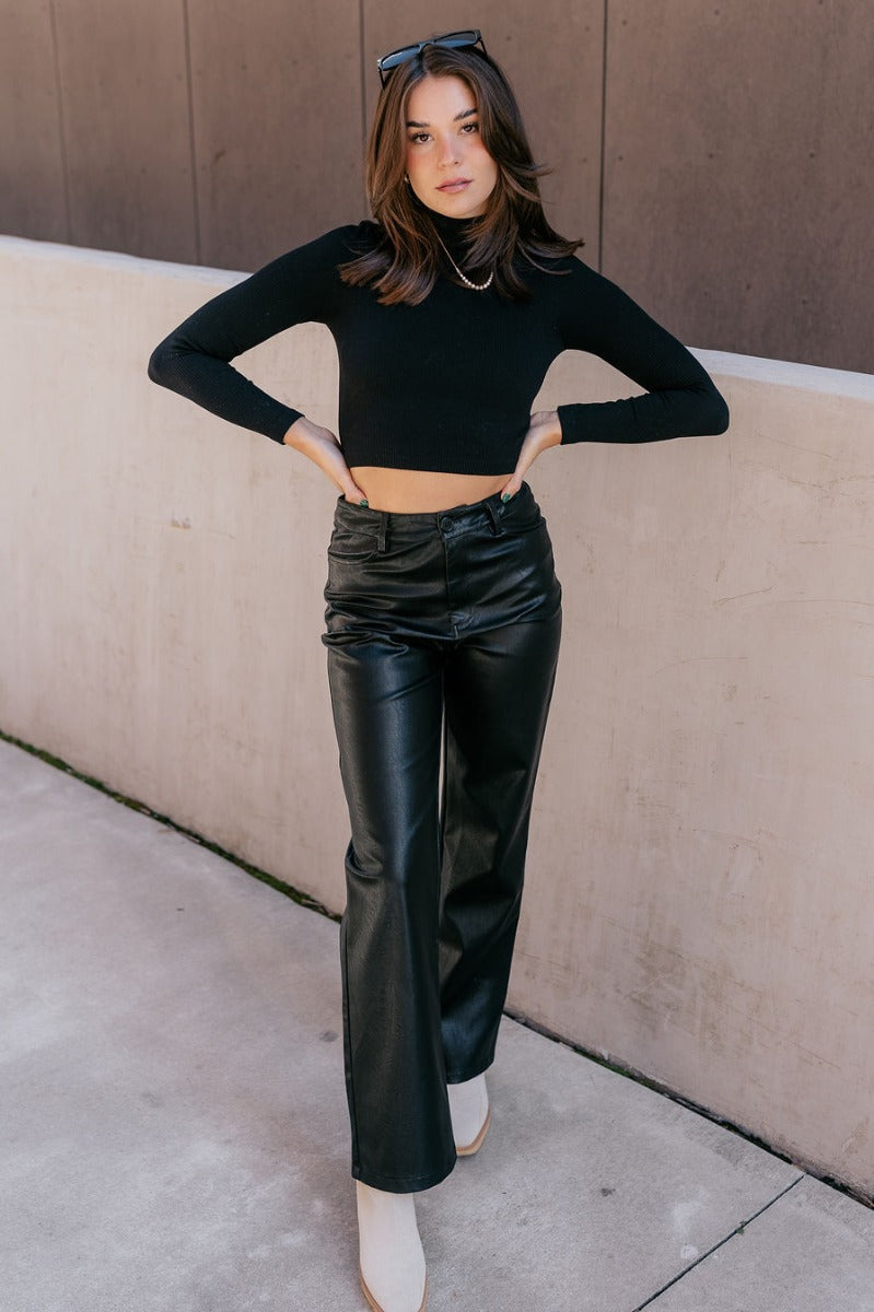 Full body view of model wearing the Naomi Black High Neck Long Sleeve Top which features black ribbed fabric, a v-neckline, and long sleeves.