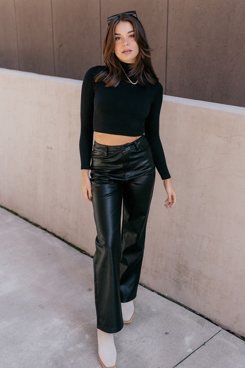 Full body view of model wearing the Naomi Black High Neck Long Sleeve Top which features black ribbed fabric, a v-neckline, and long sleeves.