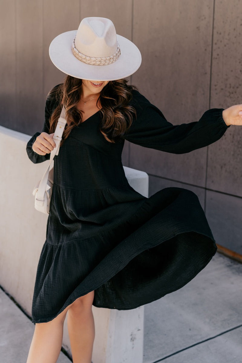 Full body view of model wearing the Wrenley Black Long Sleeve Midi Dress which features black gauze fabric, a tiered body, midi-length hem, long balloon sleeves with elastic trim, side pockets, a v neckline, and a smocked back.
