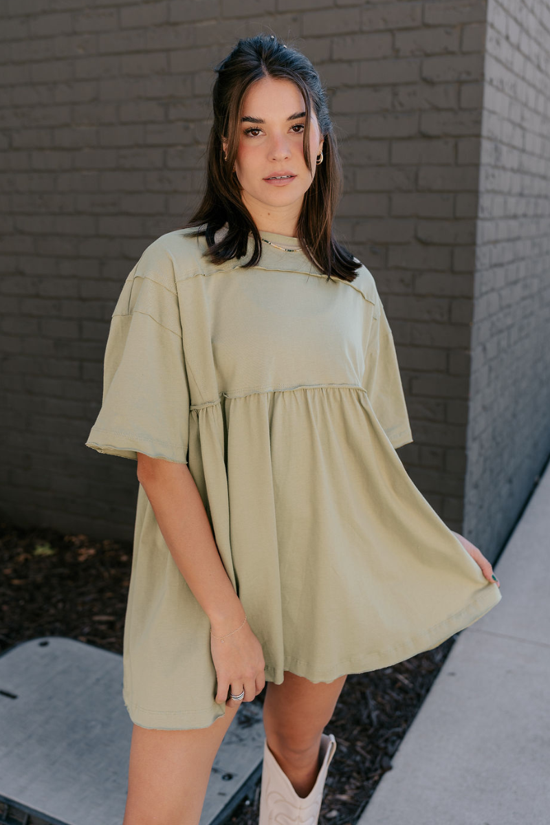 Front view of model wearing the Leah Light Olive Short Sleeve Mini Dress which features light olive knit fabric, oversized peplum body, raw hem details, round neckline and short sleeves.