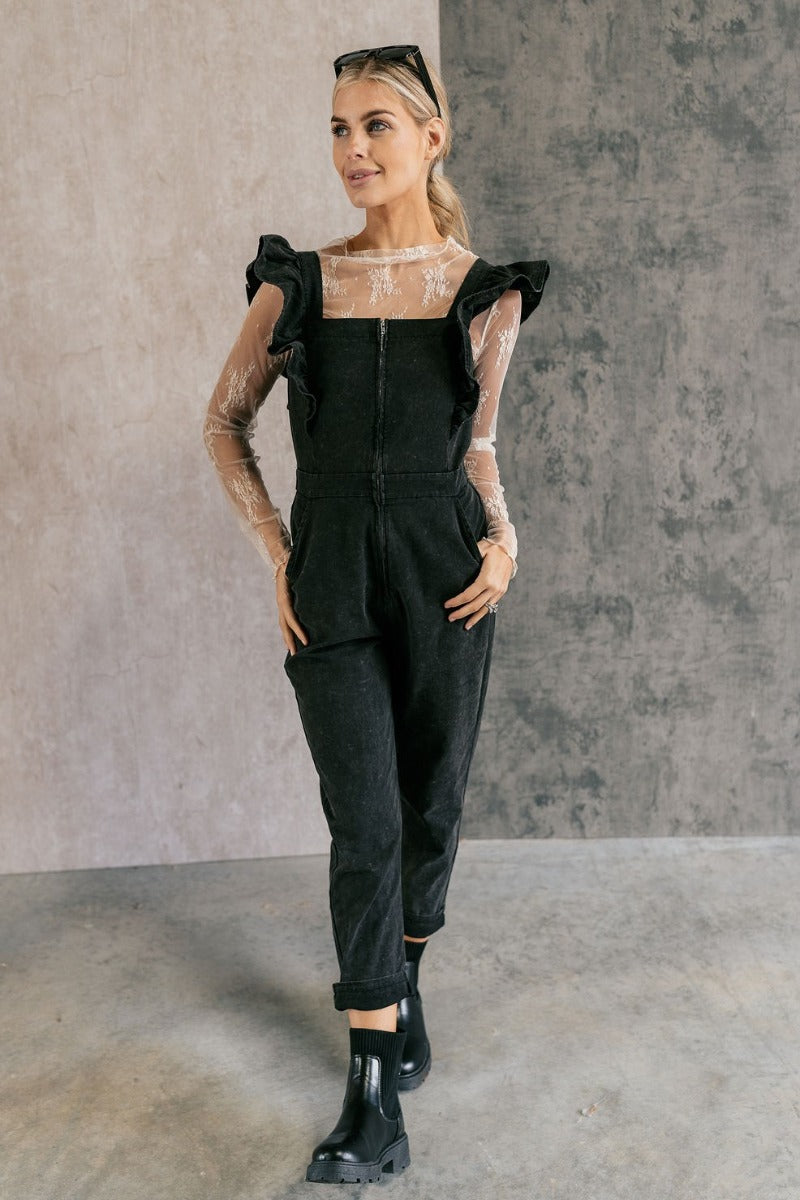 Full body front view of model wearing the Sabrina Black Denim Sleeveless Jumpsuit that has washed black denim cotton fabric, pockets, an elastic waist, a zip-up square neck, ruffle straps, and straight legs.
