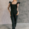 Full body front view of model wearing the Sabrina Black Denim Sleeveless Jumpsuit that has washed black denim cotton fabric, pockets, an elastic waist, a zip-up square neck, ruffle straps, and straight legs.