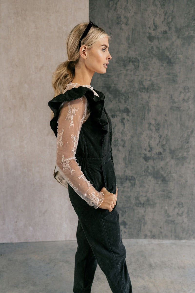 Side view of model wearing the Sabrina Black Denim Sleeveless Jumpsuit that has washed black denim cotton fabric, pockets, an elastic waist, a zip-up square neck, ruffle straps, and straight legs.