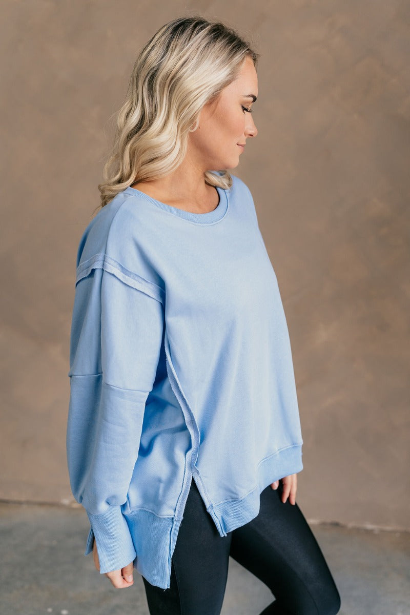 Side view of model wearing the Sadie Light Blue Long Sleeve Sweatshirt which features light blue knit fabric, distressed details, ribbed hem, slits on each side, round neckline, dropped shoulders and long balloon sleeves with cuffs.