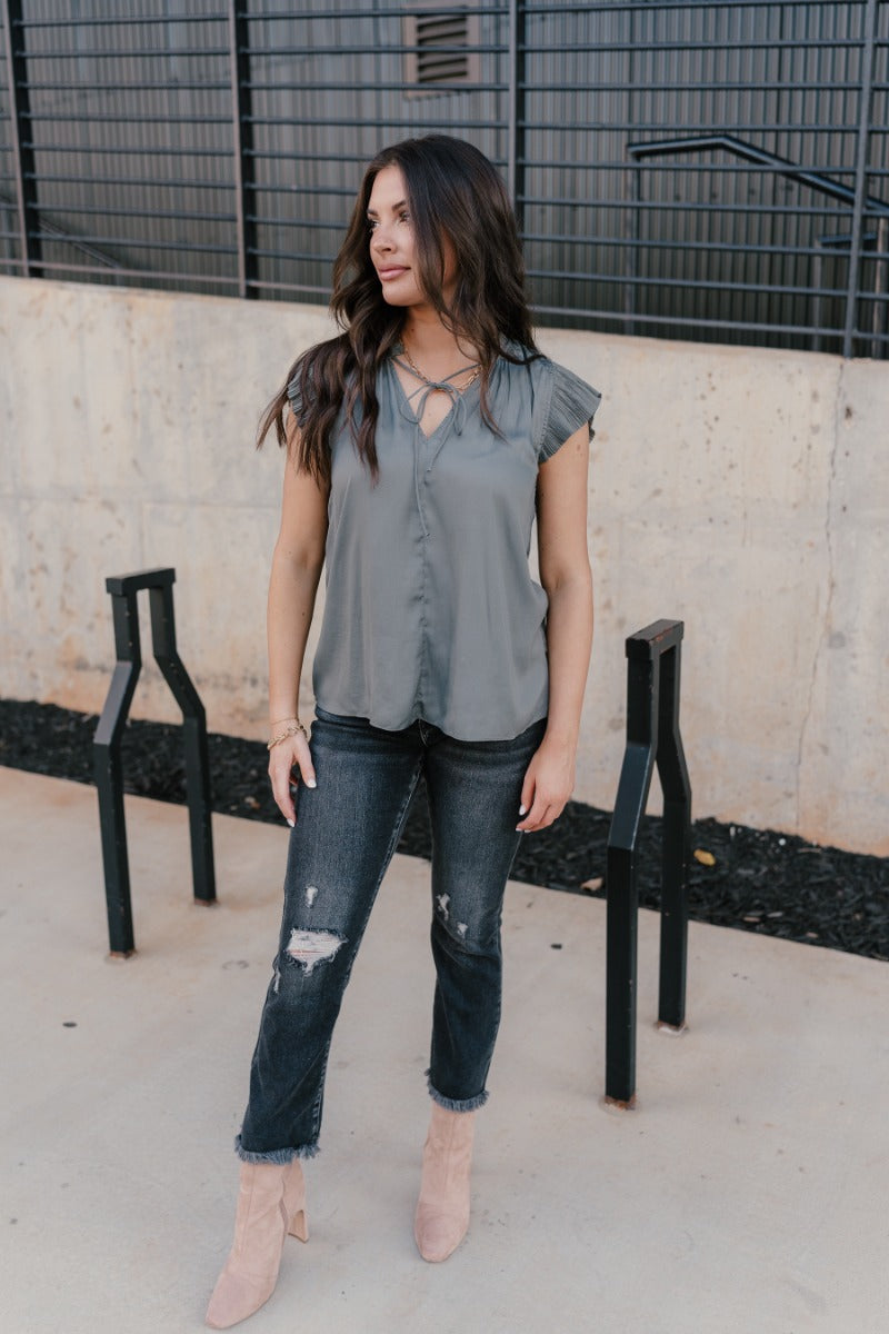 Full body front view of model wearing the Daniela Charcoal Satin Pleated Top that has charcoal grey satin fabric, a v-neck with ruffle details, an adjustable tie, and short sleeves with pleated details.