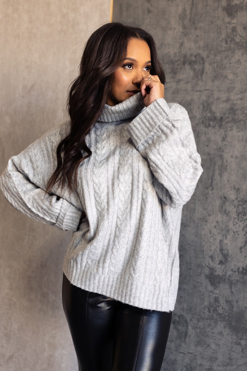 Front view of model wearing the Palmer Grey Knit Turtleneck Long Sleeve Sweater which features light grey knit fabric, a monochrome cable knit design, ribbed trim, a turtleneck neckline, dropped shoulders, and long sleeves with ribbed cuffs.