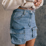 Side view of model wearing the Samira Blue Denim Mini Cargo Skirt that has blue denim fabric, mini length, pockets, belt loops and a front zipper with a button closure.