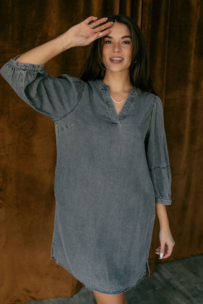 Front view of model wearing the Naomi Denim Black Half Sleeve Mini Dress which features washed black denim fabric, mini length, slight slits on each side, pockets on each side, a round neckline with a v cutout, and half balloon sleeves.