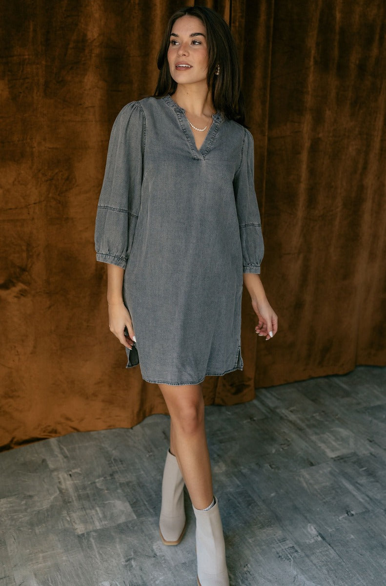 Full body view of model wearing the Naomi Denim Black Half Sleeve Mini Dress which features washed black denim fabric, mini length, slight slits on each side, pockets on each side, a round neckline with a v cutout, and half balloon sleeves.