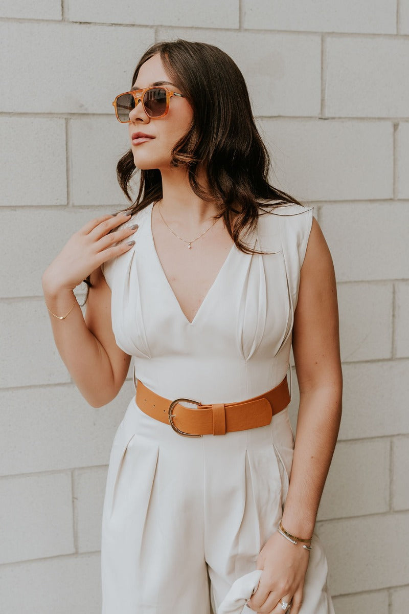 Front view of model wearing the Nova Beige Sleeveless Belted Jumpsuit which features light beige knit fabric, upper pleated details, wide pant legs, v-neckline, sleeveless, monochrome back zipper with hook closure and an adjustable camel brown belt with a